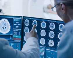 In Control Room Doctor and Radiologist Discuss Diagnosis while Watching Procedure and Monitors Showing Brain Scans Results, In the Background Patient Undergoes MRI or CT Scan Procedure.